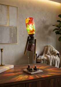 Lampe sculpturale en forme de petit personnage en bois et métal, avec une ampoule vitrail peinte à la main en guise de tête, diffusant une lumière chaude et des reflets colorés dans un intérieur.