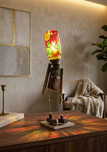 Lampe sculpturale en forme de petit personnage en bois et métal, avec une ampoule vitrail peinte à la main en guise de tête, diffusant une lumière chaude et des reflets colorés dans un intérieur.