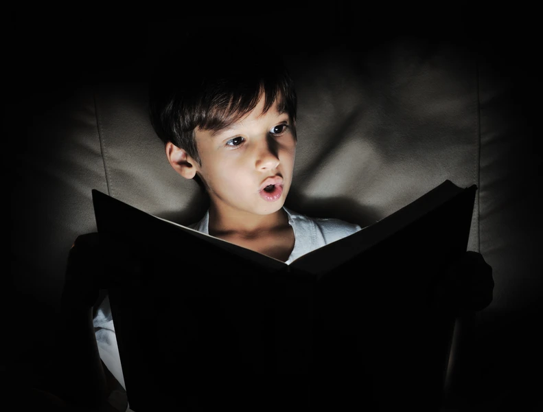 Young surprised boy sits in a dark background reading a book with light coming out of it