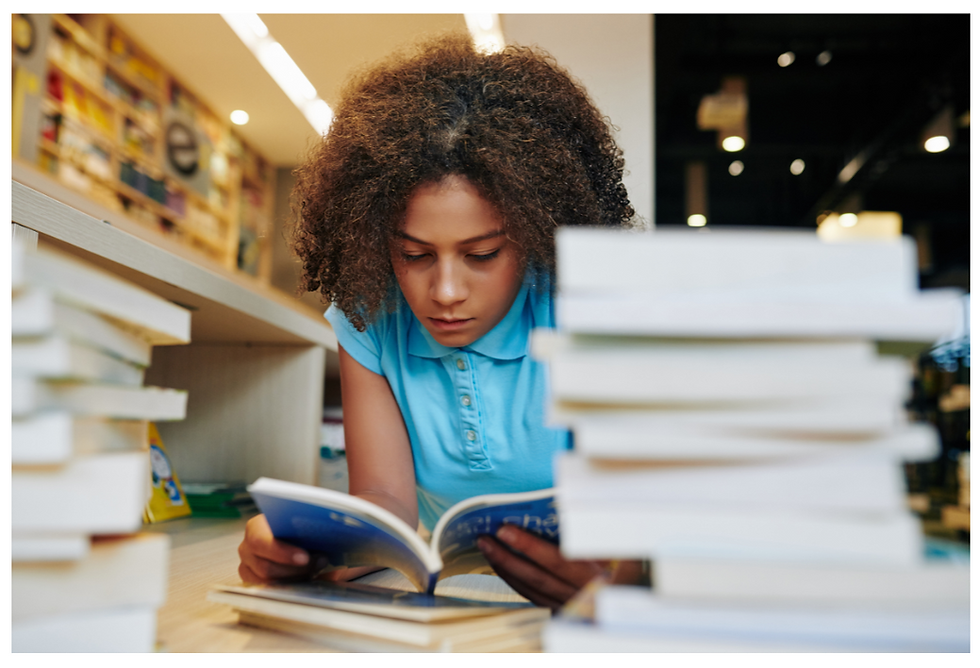Young girl in a blue shirt reads an open book between tall stacks of books in a bookstore, focused and studious.
