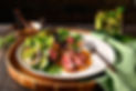 An Exclusive grass fed beef recipe image. A plate of Thai Grass Fed Beef Steak Salad with beautiful sprigs of fresh herbs and mint. The white plate sits on a bamboo platter with a rustic copper fork. There is a ramekin of sauce or dressing with a spoon in the background to the left. To the right, there is a refreshing short glass of homemade limeade with fresh mint, basil and limes.