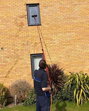 A worker uses a long pole with a brush to clean a high window on a brick building