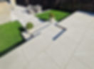 A stylish outdoor space featuring grey stone tiles, green grass, potted shrubs, and a seating area with a table and chairs