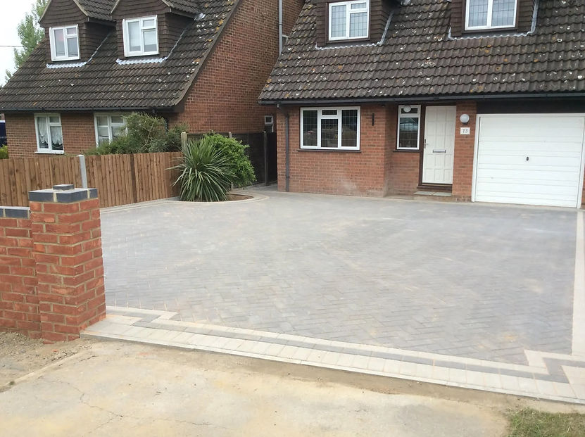 A modern brick house with a neatly paved driveway and a garage, featuring wooden fencing and greenery