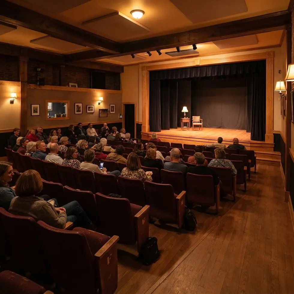 Interior photography of a small theater auditorium filled with an audience facing a stage. The atmosphere is warm, intimate, and dimly lit.