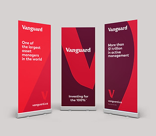 Three Vanguard branded retractable banner stands displayed as a set. Left banner reads "One of the largest asset managers in the world" on a red background. Centre banner features a deep red and maroon abstract V-shape graphic with the tagline "Investing for the 100%." Right banner reads "More than $1 trillion in active management" with the vanguard.ca URL. All banners feature the Vanguard logo and iconic V mark.