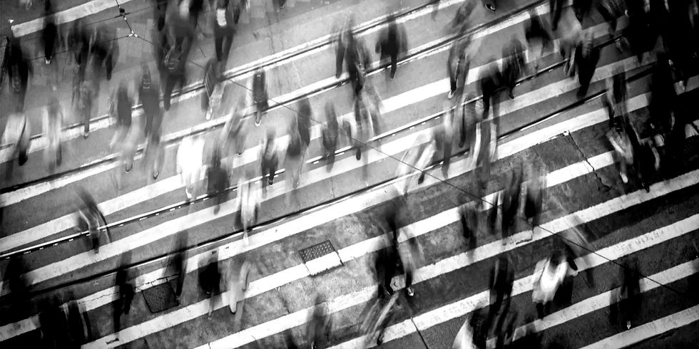 Blurred crowd crossing a street in black and white, captured from above. Striped crosswalk lines create a dynamic, bustling atmosphere.