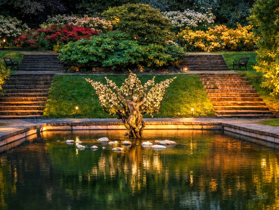 Illuminated tree sculpture in a tranquil garden pond, surrounded by steps and colorful flower bushes. Benches on each side add to the peaceful ambiance.