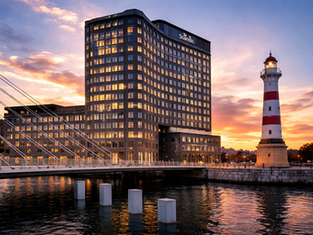 Tall modern building labeled "Scandic" and lighthouse at sunset by water. Reflections on water, warm orange sky, and a nearby bridge.