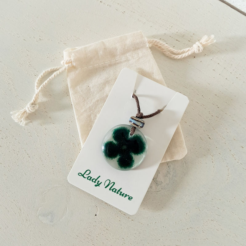 Glazen hanger van gefuseerd glas, in het glas is een groen klavertje vier gemaakt van glas. Boven aan de hanger zit een grijs