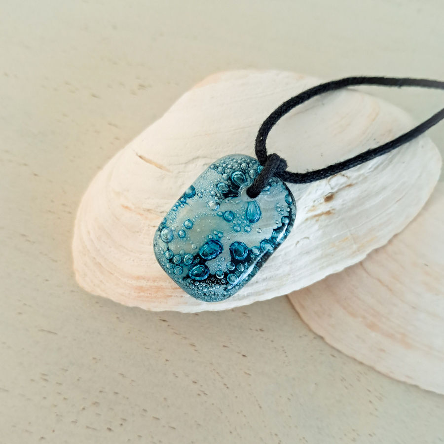 Hanger van gefuseerd glas, in het glas zitten aqua blauwe bubbels. De hanger zit aan een zwart waxkoord.
