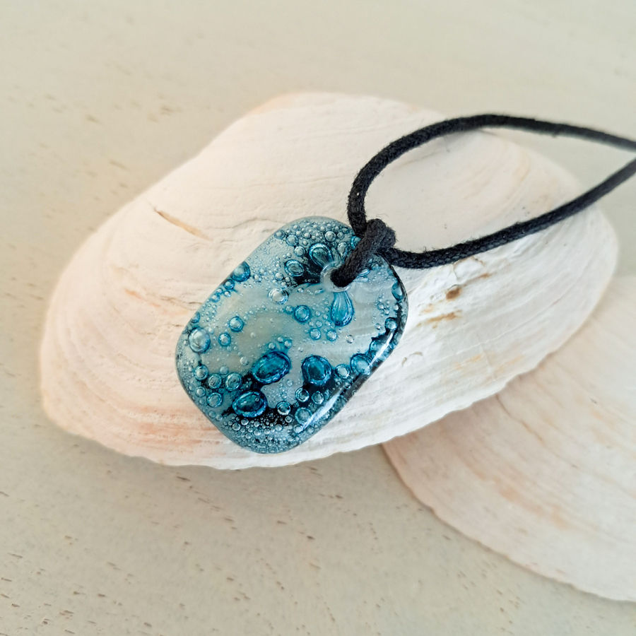 Hanger van gefuseerd glas, in het glas zitten aqua blauwe bubbels. De hanger zit aan een zwart waxkoord.