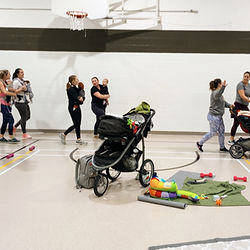 Stroller Yoga Fitness class Winnipeg. A group of moms and babies are walking around a gym, being social, and talking.