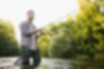 Angler in waders fishing a river in Arizona Hunt Unit 5B North, surrounded by green trees—serene Anderson Mesa setting for trout fishing and outdoor recreation.