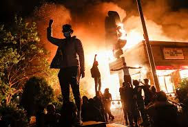 Protesters stand in front of a burning building during a nighttime demonstration in the United States.