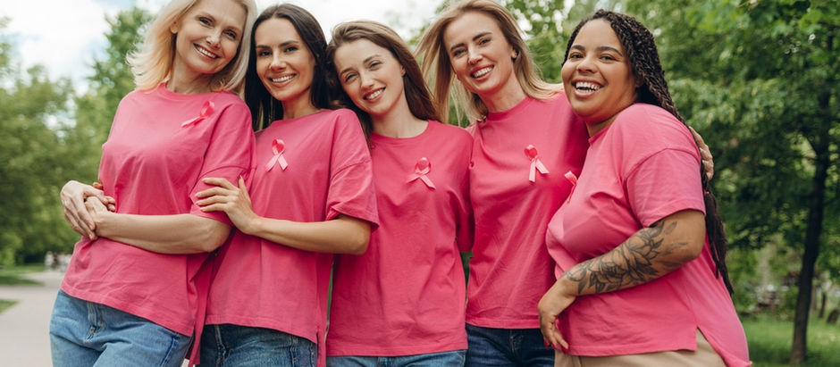 Cinco mulheres com blusa rosa e laço, símbolo do outubro rosa, se abraçam em um parque.
