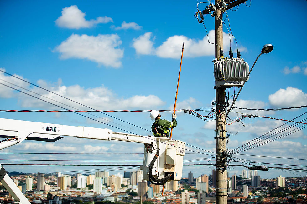 Em um guindaste apropriado para manutenção da rede elétrica, técnico da Neoenergia está fazendo a manutenção de uma rede elétrica com céu no fundo e também prédios altos.