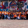foto registrando encontro do grupo Elas com Elas, com várias mulheres de camisa laranja olhando para a fotografia. O fundo é composto por edificação colorida em grafite.