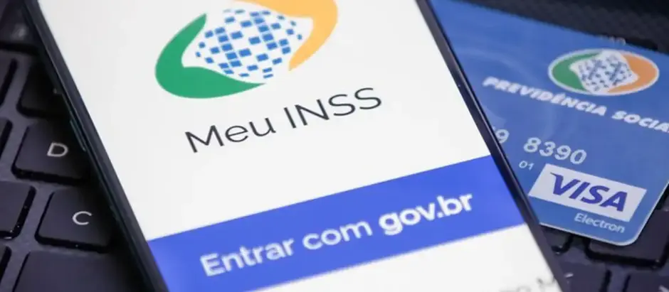 Em uma tela de celular, está a logo do aplicativo MEU INSS, com cartão do INSS de fundo e teclado do computador por baixo