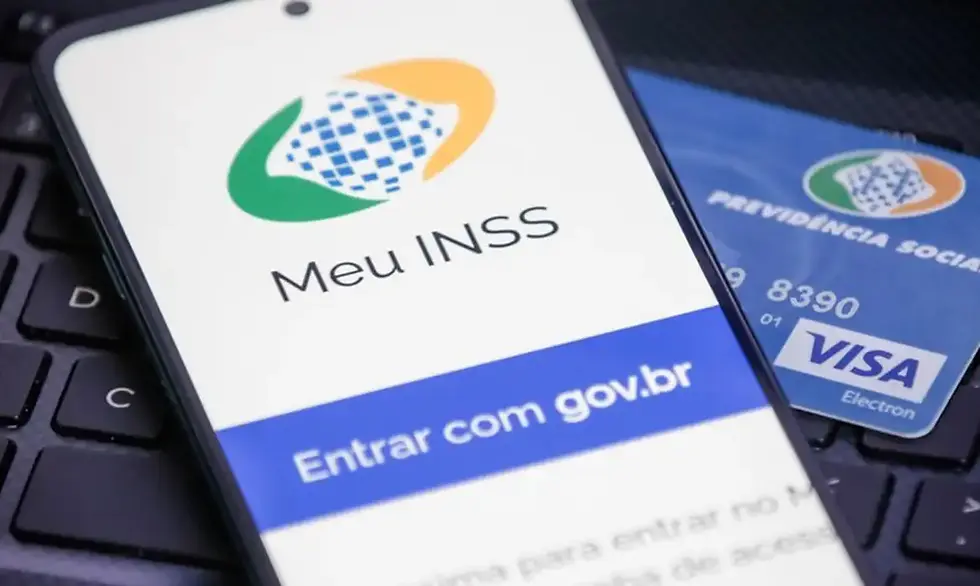 Em uma tela de celular, está a logo do aplicativo MEU INSS, com cartão do INSS de fundo e teclado do computador por baixo
