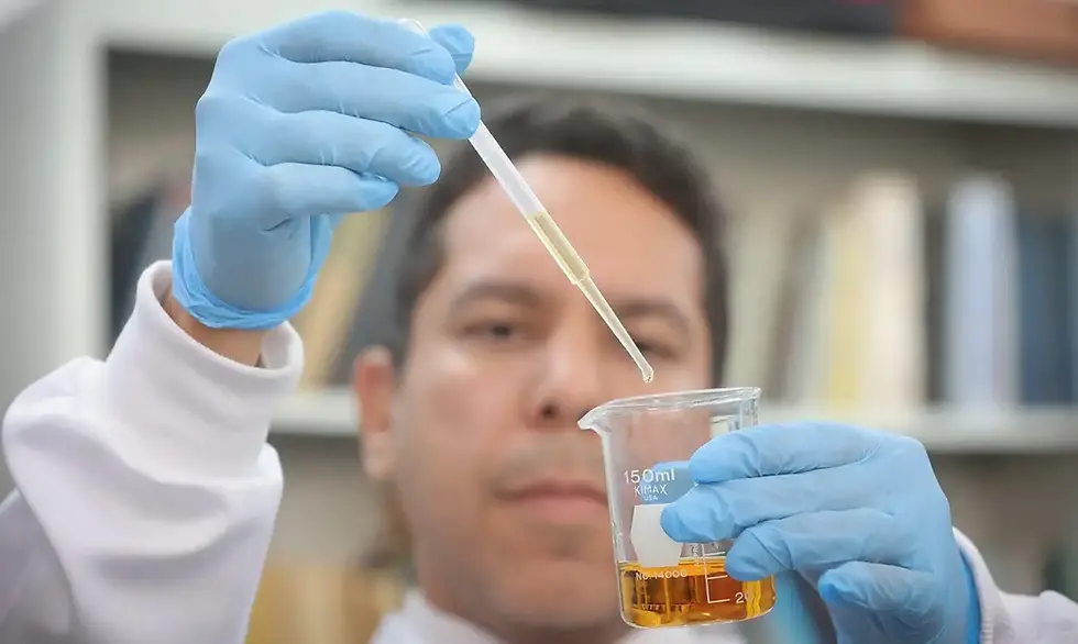 A imagem mostra uma pessoa usando luvas azuis de proteção, segurando um béquer de 150 ml com um líquido amarelado e uma pipeta com uma pequena quantidade do mesmo líquido. O fundo parece ser um laboratório, com prateleiras e livros desfocados.