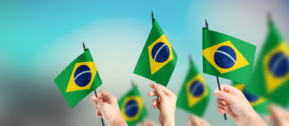 A imagem mostra várias mãos de pessoas segurando pequenas bandeiras do Brasil (bandeiras verde-amarela com o globo azul no centro e a faixa branca com "ORDEM E PROGRESSO") erguidas para o alto. O fundo é um céu azul claro e limpo, com um leve efeito de luz ou brilho no centro superior, dando uma sensação de patriotismo, união e celebração. As bandeiras estão em foco nítido no primeiro plano, com algumas ligeiramente desfocadas ao fundo, criando profundidade. 