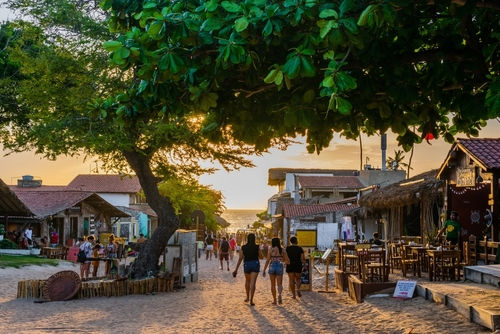 Rua de areia e casas rústicas com vista do mar. Há turistas caminhando em direção à praia no nascer do sol.