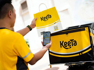 Imagem de um entregador da Keeta segurando uma sacola e um celular, mostrando a interação com o aplicativo de entrega. A caixa de entrega também é da Keeta, destacando a marca em um ambiente urbano.