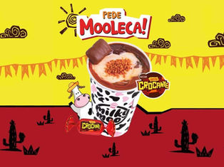 Imagem de uma sobremesa de mooca com calda de chocolate e enroladinho de biscoito, apresentada em um cenário com tema de fazenda e elementos divertidos, promovendo a campanha Peça Mooca.