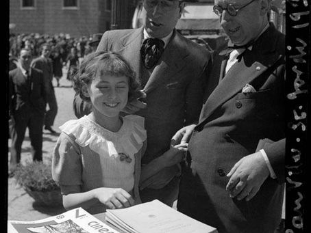 Black-and-white photo of a young girl smiling at a table covered with books and papers, standing beside two men in suits and hats at an outdoor book stall, with a small crowd and buildings in the background.