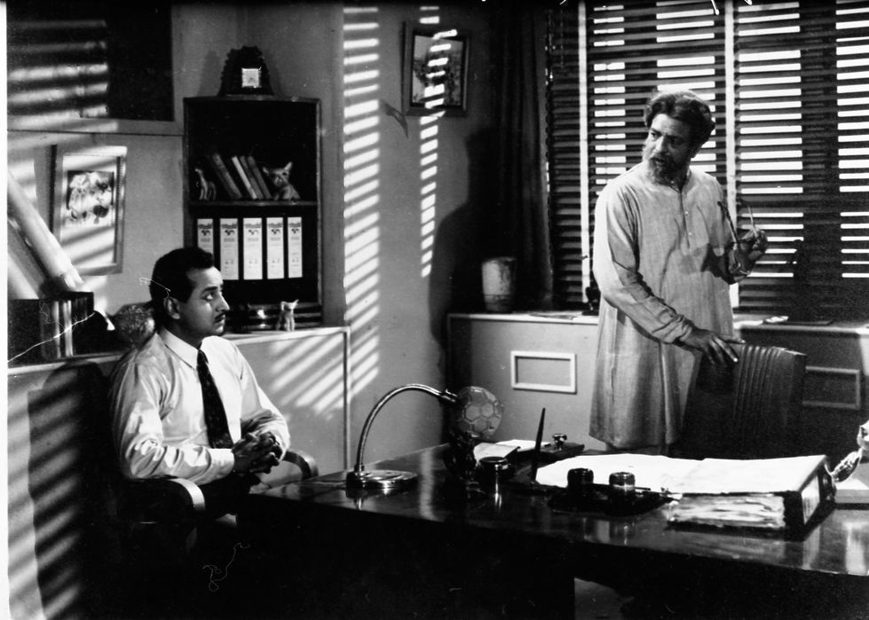 a man sits at a desk in front of a window while another man stands behind him