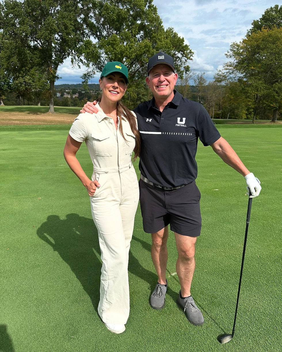 Smiling man and woman pose on a golf course, arm in arm, with a club; his shirt reads U Football.
