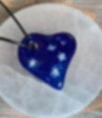 The image shows a deep blue glass heart necklace. It sits on a round piece of white rock with a leather cord attached to the heart.  Embedded in the heart are little white stars to represent the little bit of light in the dark of the night.