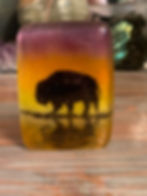 An image of a rectangle glass art piece.  It shows the silhouette of a buffalo agent a colorful sunset in pinks, purples, oranges and yellows.  Its reflection in water is the bottom third of the small piece.  It stands on it's own on the  table surface.