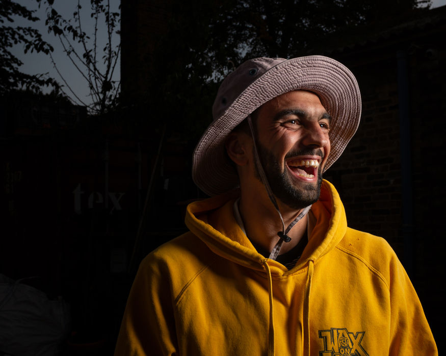 Man in yellow hoodie and wide-brimmed hat smiling in golden sunlight against dark foliage, photographed by Aemilia Cumberland