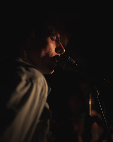 Performer singing into microphone in dramatic side lighting against black background, photographed by Aemilia Cumberland