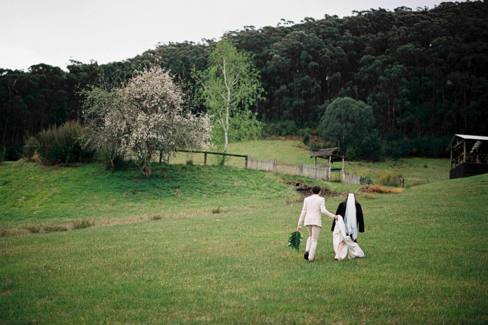 groom holds up brides dress as they walk through a grassy field