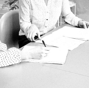 Two women in polka-dot blouses review and write on papers at a conference table in a black-and-white office scene