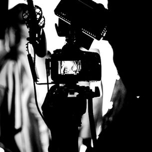 Silhouetted video camera and microphone on a set, with a blurred person in the background, creating a stark black-and-white scene.