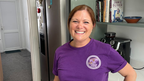 Emily in a kitchen wears a purple Big Purple Pants T-shirt, with books and a coffee machine behind her.