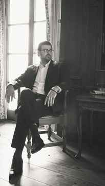 Man in suit sits by window, wearing glasses, engagement photography Chicago.
