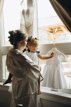 Woman holding a child looking at a white dress near a window; wedding photography Chicago.