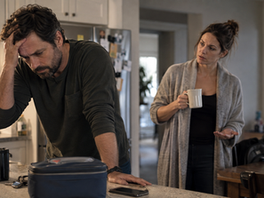 Tense man leaning on a kitchen counter with his hand on his forehead while his partner gestures nearby.
