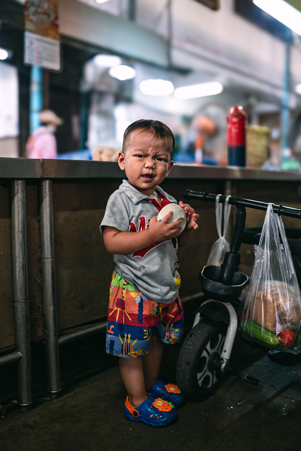 Curious Thai toddler in market stall holding a bowl beside balance bike and groceries – candid stree