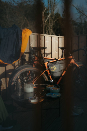 Orange bikepacking bike with Apidura bags seen through a tent entry, breakfast setup with kettle and bowls visible in warm light, Vestkystruten, 2022