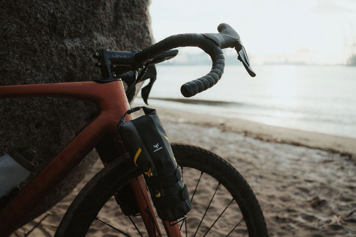 Apidura Expedition Cargo Cage Pack mounted on orange gravel bike fork on sandy Elbe shore, Hamburg harbor in background, December 2022