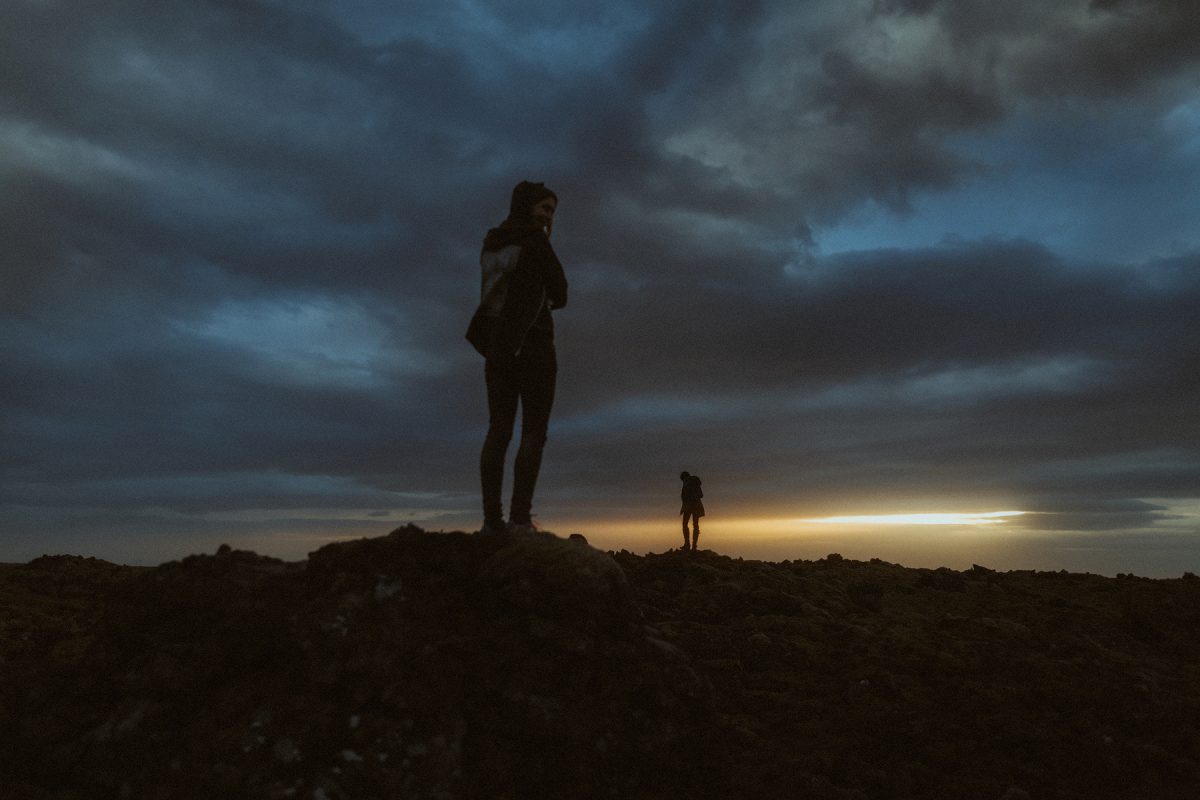 Silhouettes of two people standing on rocky terrain under a dramatic, cloudy sky at dusk.