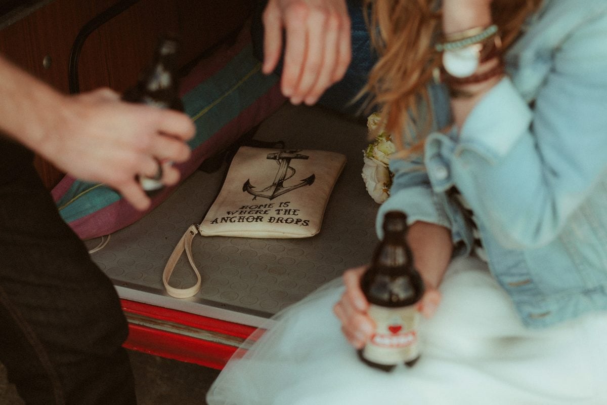 Close-up of a just-married couple sharing a beer in the back of their van during their St. Peter-Ording elopement. A canvas pouch reading “Home is where the anchor drops” lies beside them—surrounded by sandy feet, denim jackets, and post-ceremony calm.