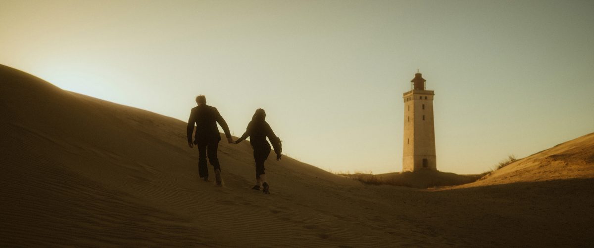 Elopement at Rubjerg Knude, Denmark: A couple walks hand in hand through the windswept dunes at sunset, the iconic lighthouse in the distance. Raw, cinematic, and full of quiet connection—perfect for a wild and intimate wedding at the North Sea.