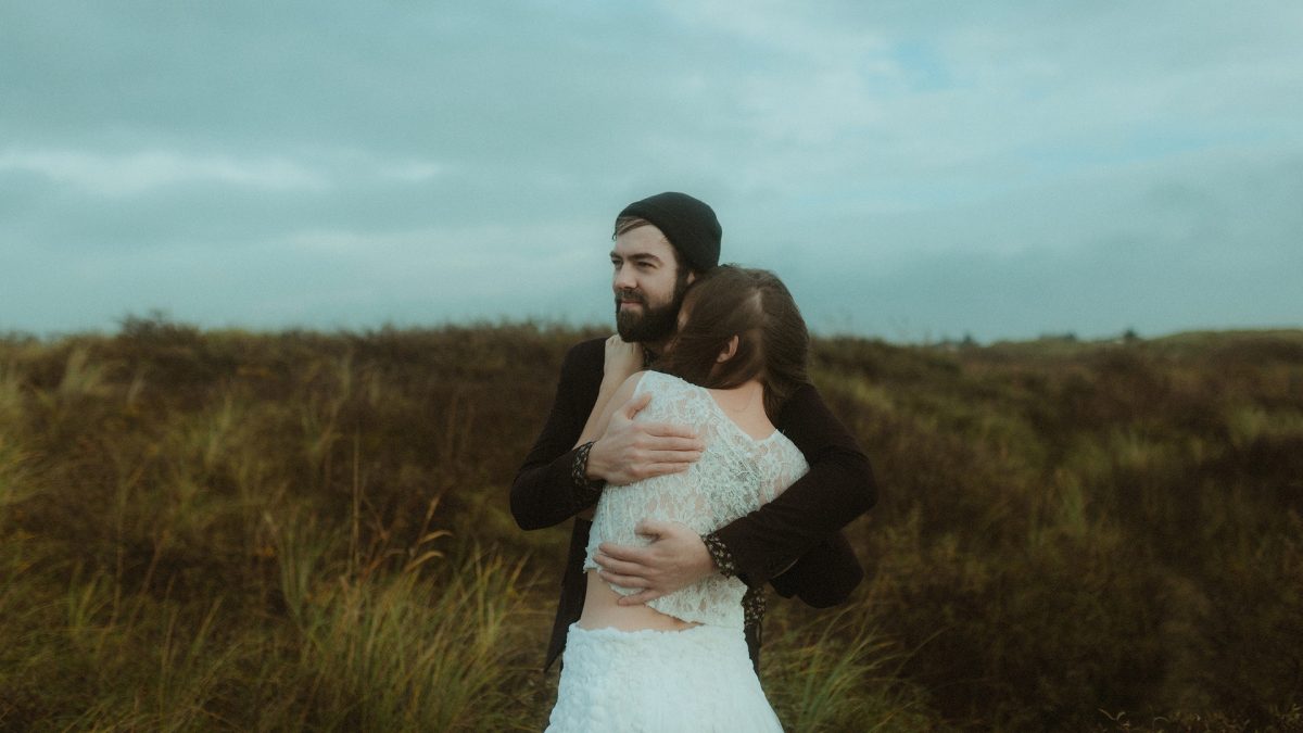 Couple embracing deeply in the wild dunes of Rømø, Denmark—her wedding dress softly blending into the wind-swept grass, his arms holding her close as the sky hangs heavy with soft, moody clouds. A quiet, raw moment of connection in nature.